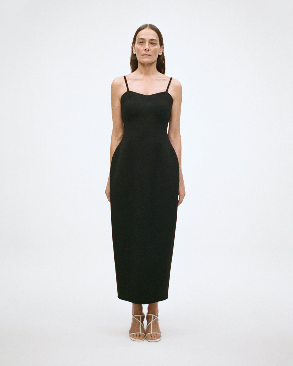 Woman wearing a black ankle lentgth, sculptural double faced wool dress with spagetti straps on a white background