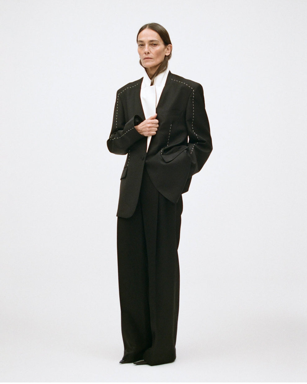 Woman wearing a black suit with white lapels and a contrasting stitch down the sleeves on a white background