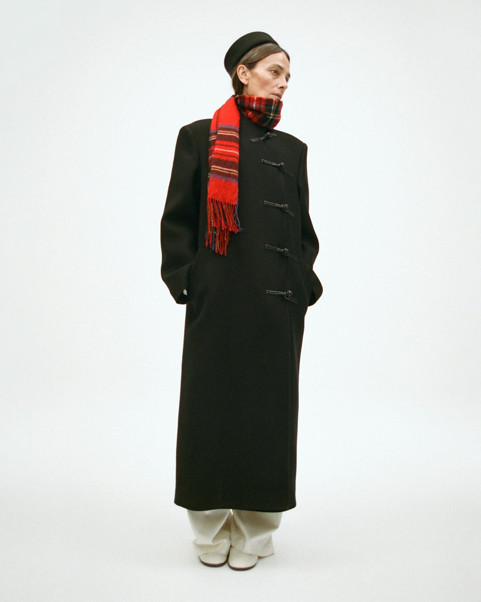 Woman wearing a pillbox hat, a long black coat, white pants, and a red scarf with a plaid pattern on a white background