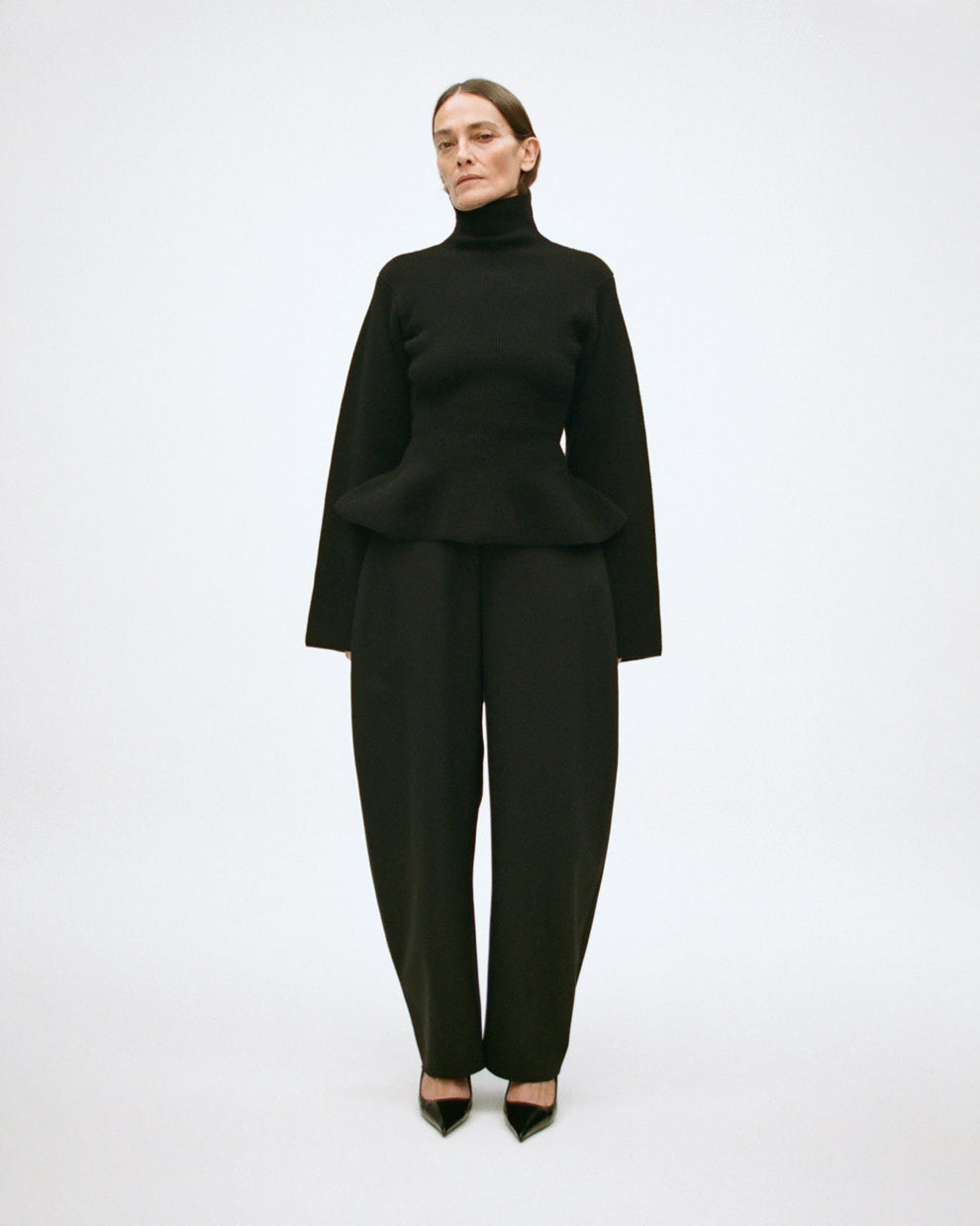 Woman wearing a black turtleneck sweater with peplum bottom, and black barrel leg pants on a white background