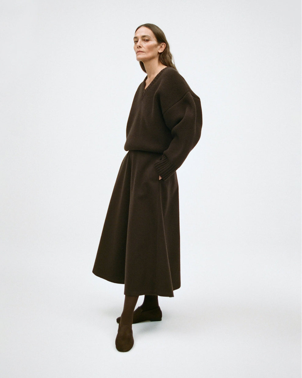 Woman wearing a dark brown thick v-neck sweater with a dark brown wool a line skirt on a white background