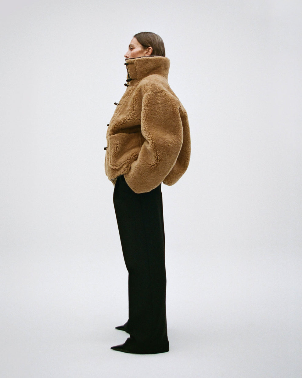 Woman wearing a brown shearling jacket with funnel collar and black pants on a white background