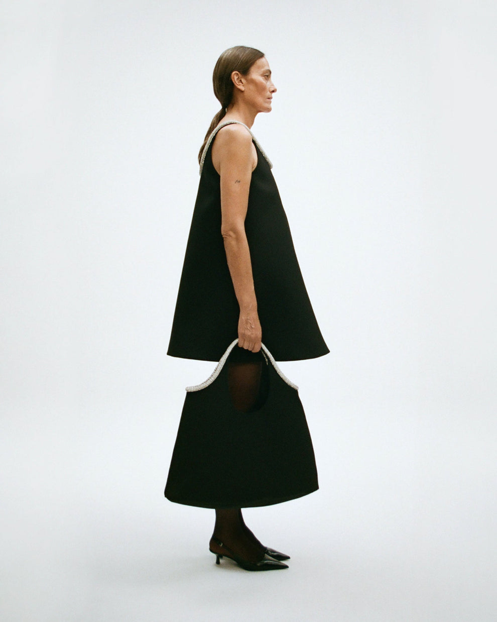 Woman wearing a black sleeveless a-line short dress while holding a black tote bag on a white background