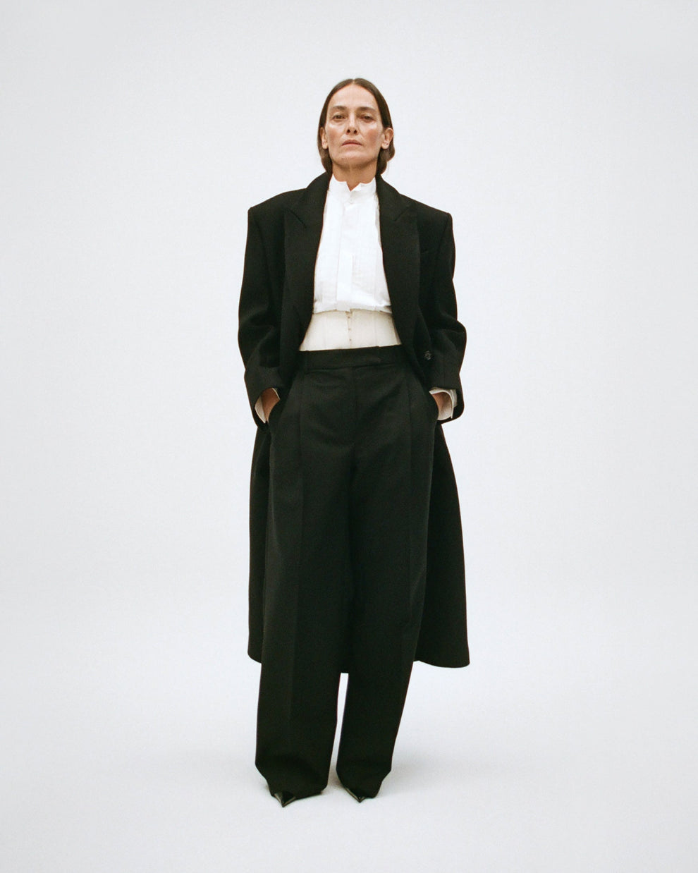 Woman wearing a black coat with a white shirt an black trousers with a cream corset detail on a white background