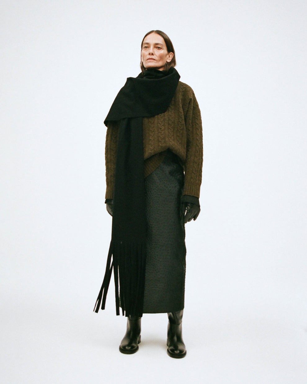 Woman wearing a forest green sweater, black long scarf with fringes, and skirt with ostrich texture on a white background