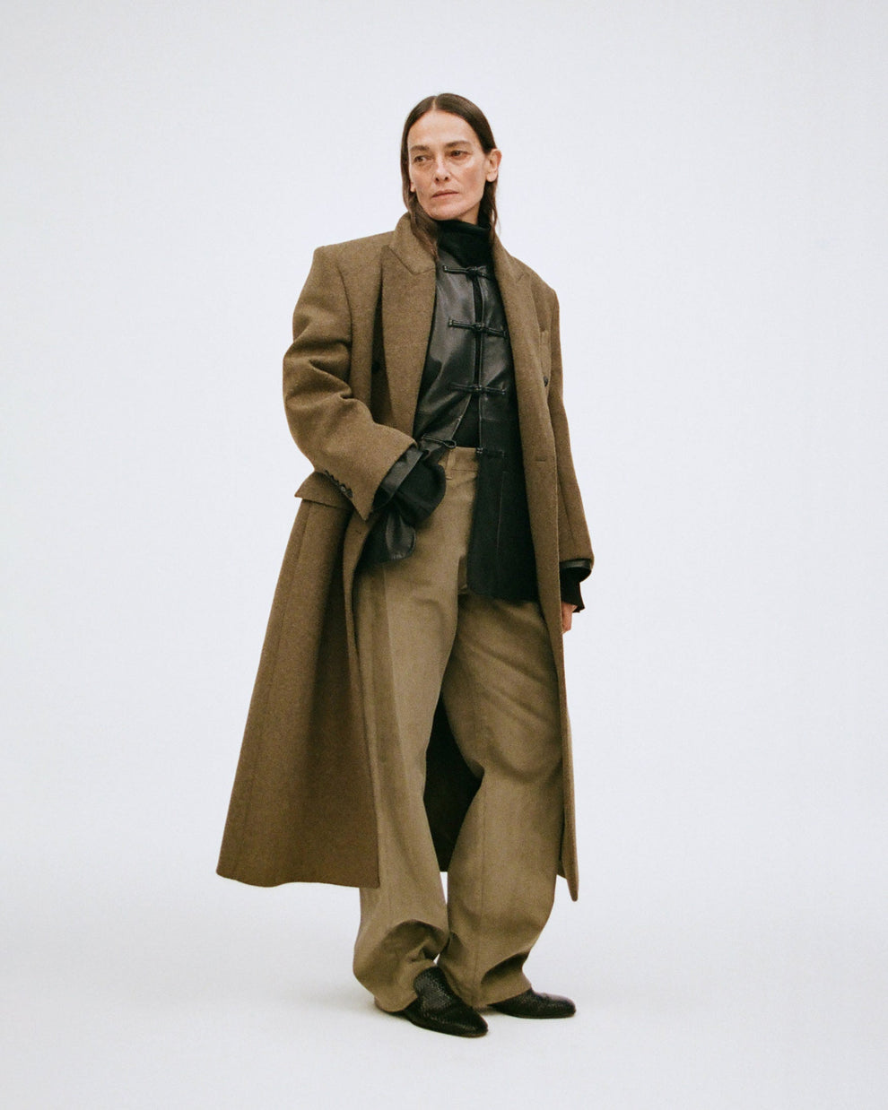 Woman wearing a long forest green wool coat, with a black leather jacket underneath, and sage colored corduroy trousers on a white background