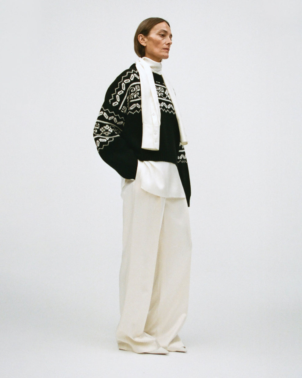 Woman wearing a black pullover with a white fair isle print, over a cream turtleneck shirt with and cream satin pants on a white background