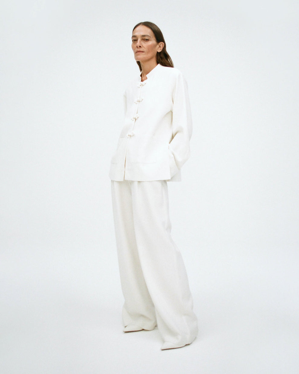 Woman wearing a white outfit consisting of a jacket with mandarin closure and a wide leg white trouser, on a plain background